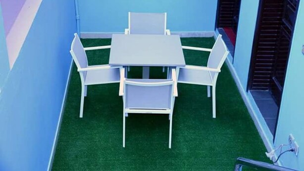 Un pequeño espacio al aire libre con una mesa y cuatro sillas, rodeado de paredes azules y césped artificial verde.