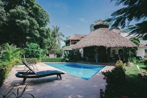 Un área de piscina con una estructura de techo de paja, sillones de descanso y exuberante follaje.
