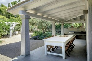 Eine Terrasse im Freien mit weißem Tisch und Stühlen, einer Pergola darüber und einem Grillplatz.