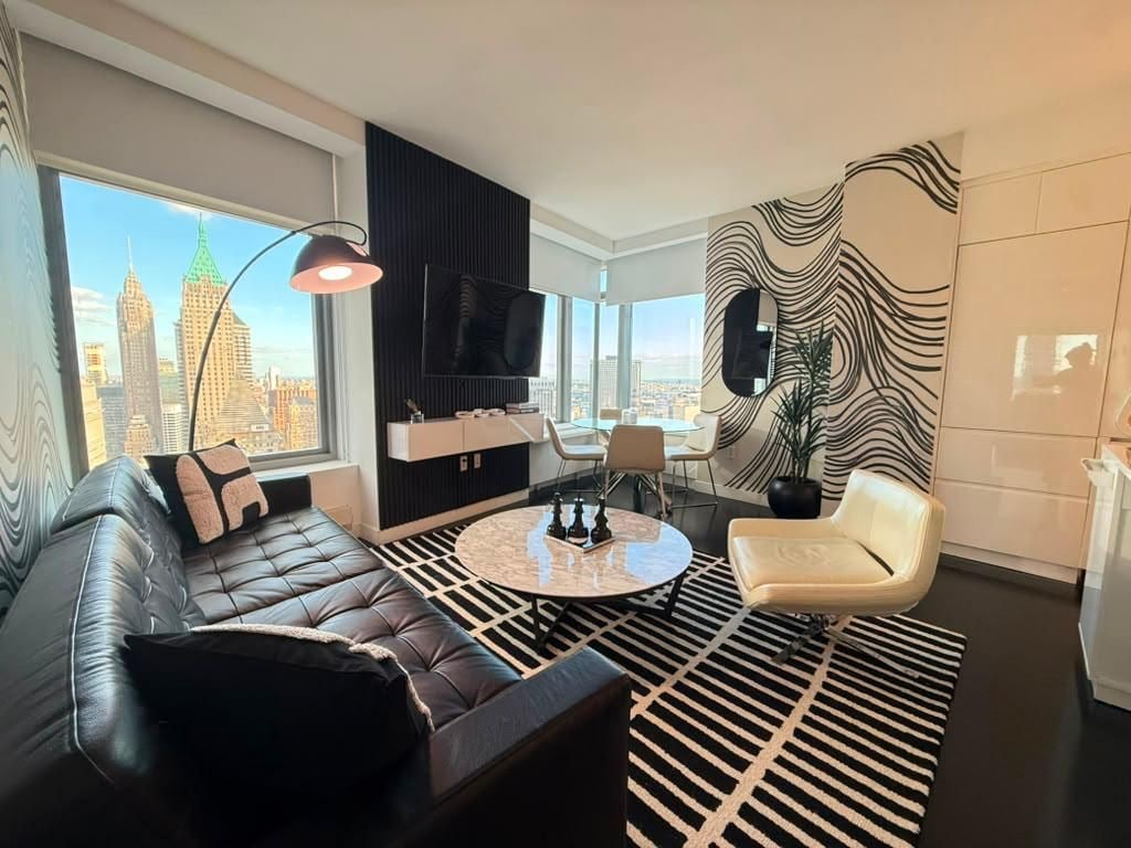 Un salon moderne avec un canapé en cuir, une table basse ronde et un fauteuil blanc. Une grande fenêtre offre une vue sur les bâtiments de la ville.