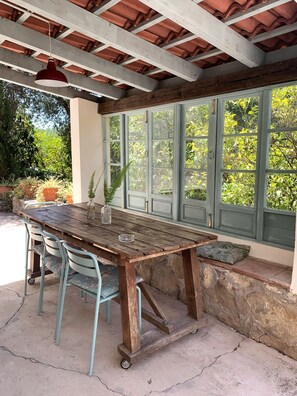 A rustic wooden table with chairs, a hanging pendant light, and a view through green-framed windows.