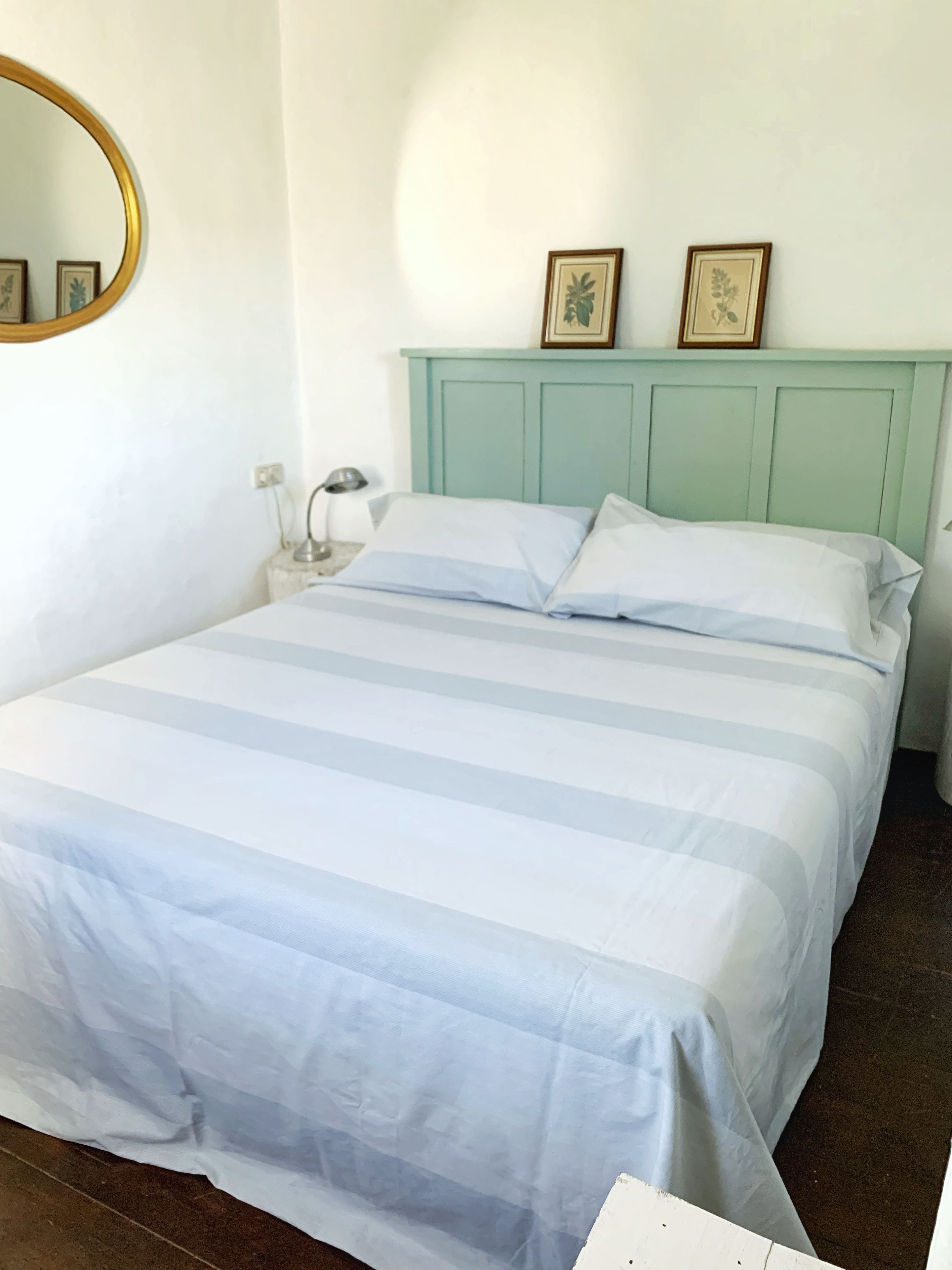 Una cama con colcha a rayas, una lámpara de noche y un espejo instalado en la pared.