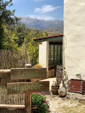 A rustic outdoor area with a wooden sign reading 'Casa Alagrolo,' a small structure with a window, and a view of mountains in the background.