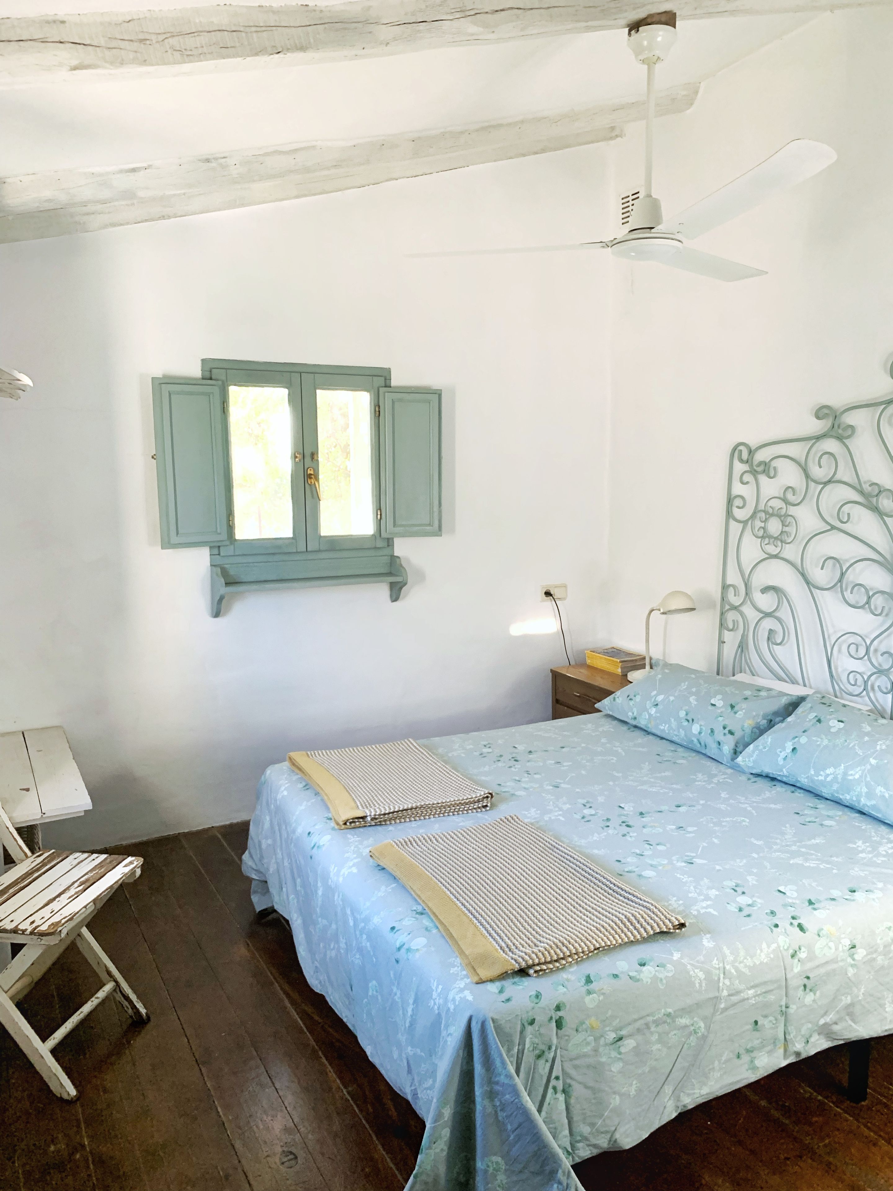 A bedroom with a bed, a window with shutters, a ceiling fan, and a bedside table with a lamp.
