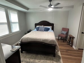 Een slaapkamer met een bed, een stoel, een nachtkastje, een plafondventilator en een raam met jaloezieën.