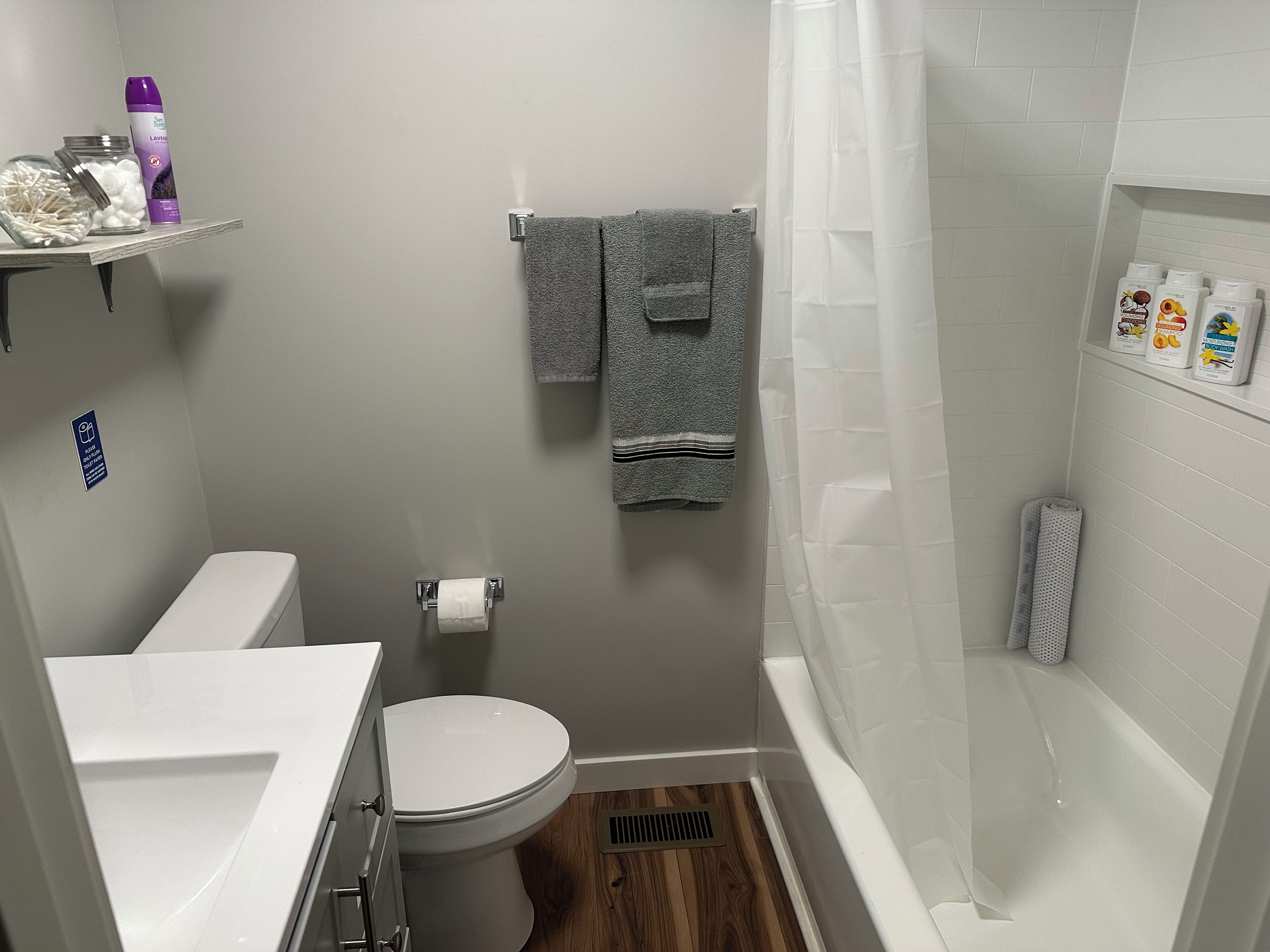 A compact bathroom with a toilet, sink, shower, and a small shelf with toiletries.
