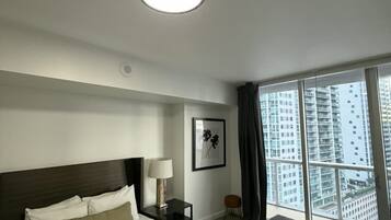 A hotel room with a large bed, a nightstand, a lamp, a framed picture, a small stool, and a view of tall buildings through a large window.