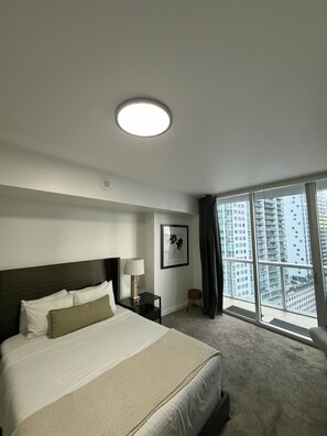 Habitación de hotel con una cama grande, una mesita de noche, una lámpara, un cuadro enmarcado, un taburete pequeño y vistas de edificios altos a través de un ventanal amplio.