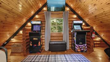 Un acogedor cuarto ático con paredes de madera, un ventanal grande y dos máquinas de videojuegos.