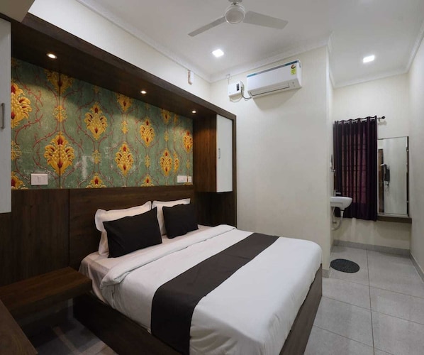 A hotel room with a bed, a headboard with a patterned wallpaper, a ceiling fan, an air conditioning unit, and a bathroom with a sink and a mirror. - Classic Djr Grand (Puducherry)