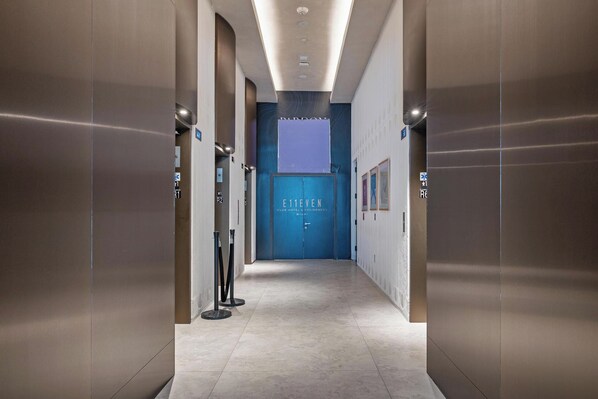 Un couloir avec des ascenseurs, une porte bleue portant l’inscription « E11EVEN CLUB HOTEL & RESIDENCES MIAMI », et un sol carrelé.