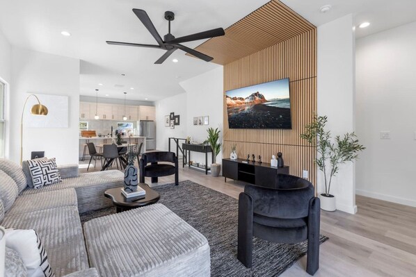 Uma sala de estar moderna com um conjunto de sofás, uma mesa de centro, uma poltrona preta, uma TV fixada em um painel de madeira e um ventilador de teto.