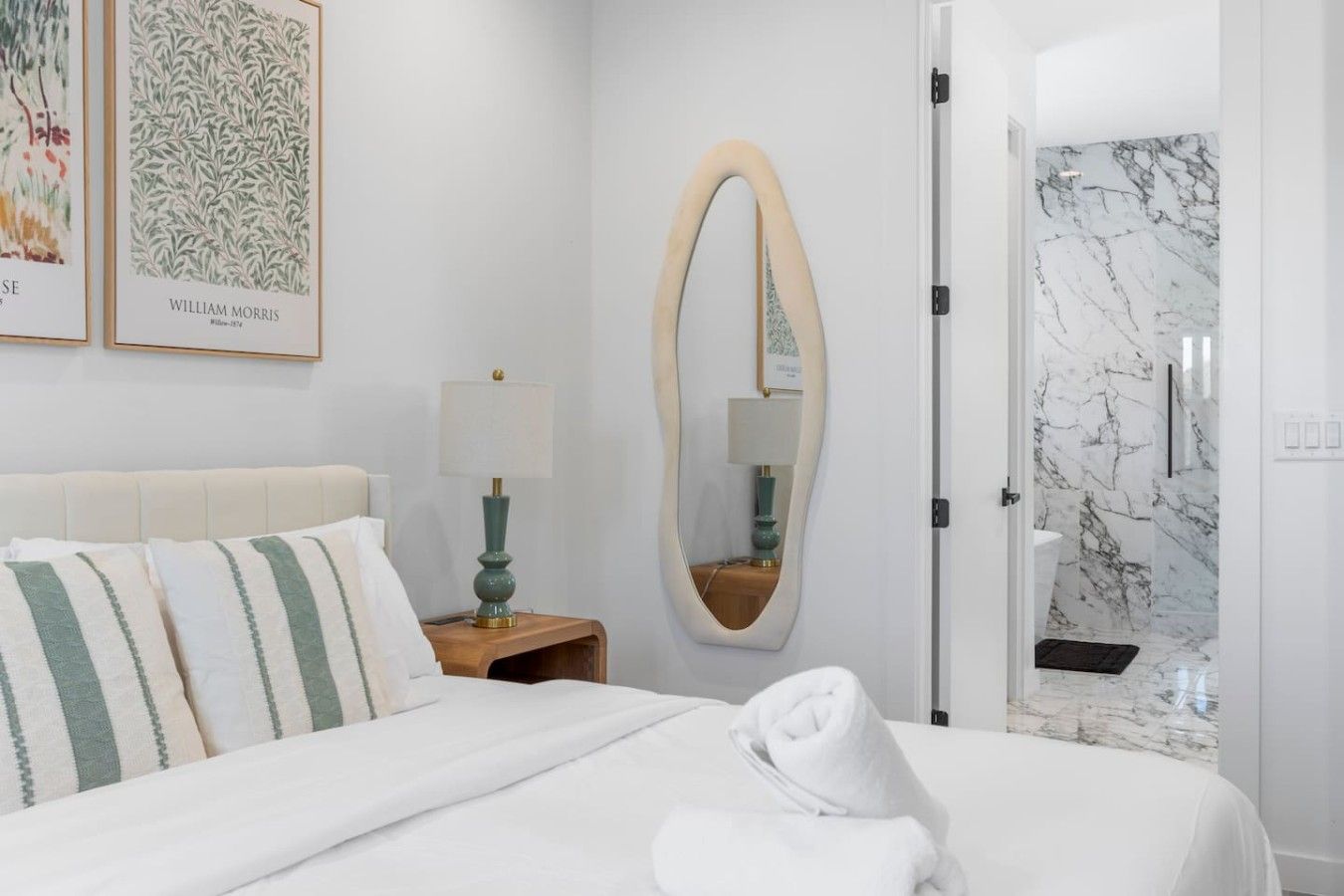 Una habitación de hotel con una cama, una mesita de noche con lámpara, un espejo y una puerta que da a un baño con ducha de mármol.