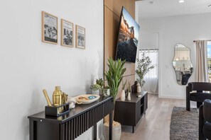 Ein modernes Wohnzimmer mit einem schwarzen Konsolentisch, einem flachbildfernseher an der Wand, einem Essbereich mit einem schwarzen Stuhl und einer durch eine Glastür sichtbaren Küche.