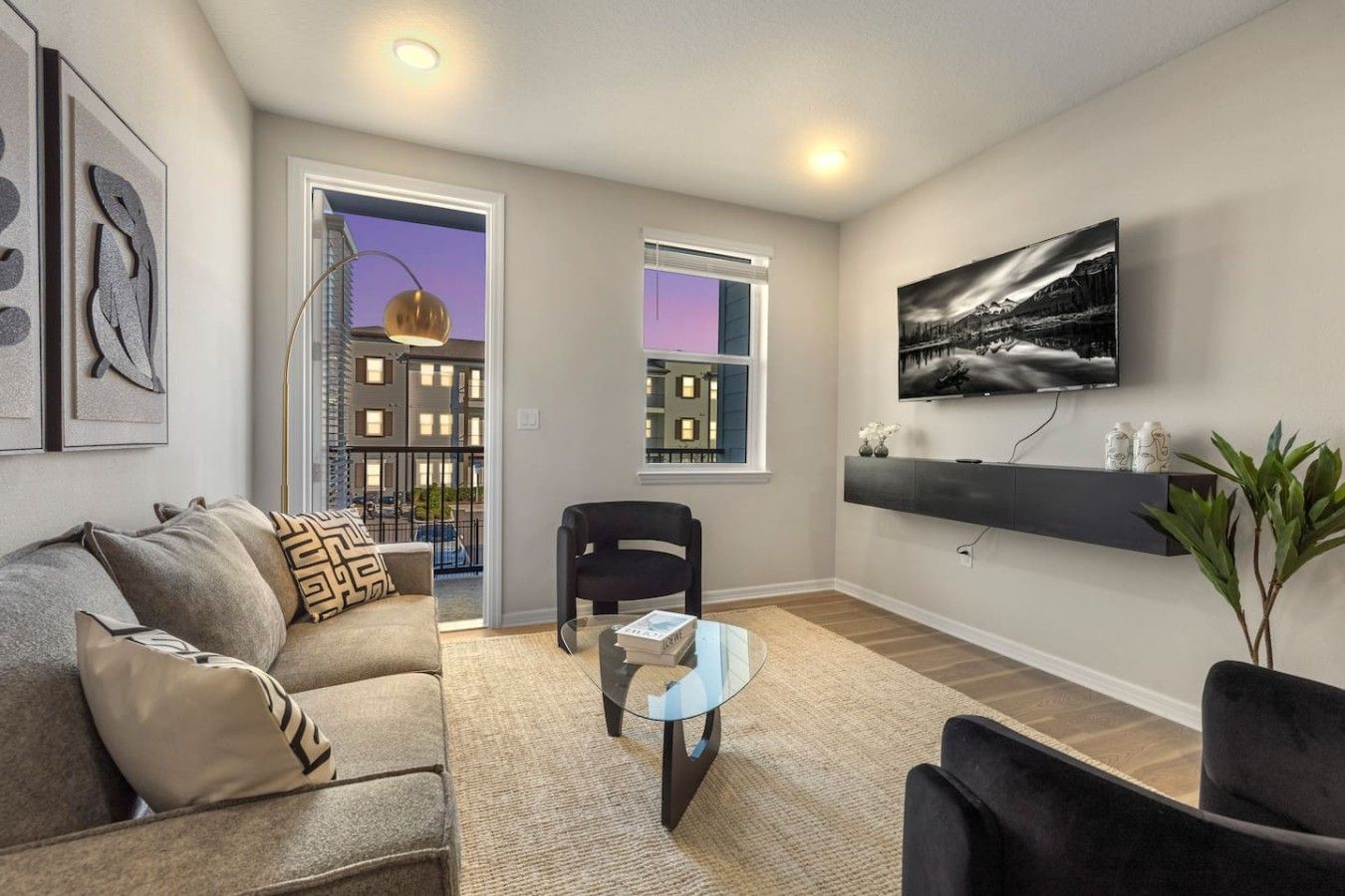 A modern living room with a sofa, a coffee table, a TV mounted on the wall, and a view of buildings through the windows.