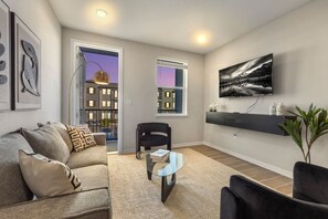 A modern living room with a sofa, a coffee table, a TV mounted on the wall, and a view of buildings through the windows. - Spacious Largo Escape | Poolside 2BR Stay (Largo)