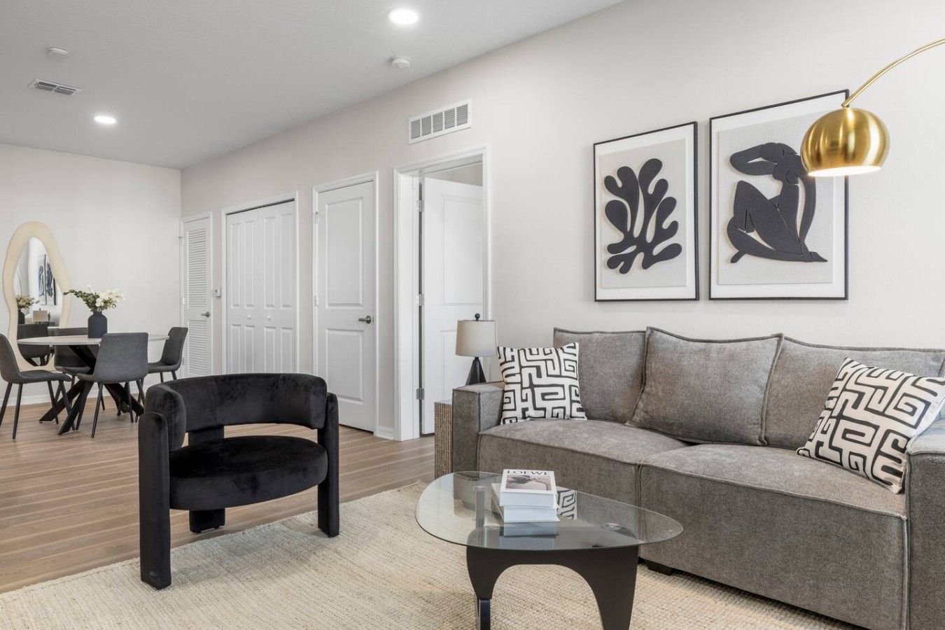 A modern living room with a black armchair, a grey sofa, and a glass coffee table. There are two framed abstract art pieces on the wall, a lamp, and a dining area with a table and chairs.