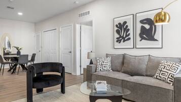 A modern living room with a black armchair, a grey sofa, and a glass coffee table. There are two framed abstract art pieces on the wall, a lamp, and a dining area with a table and chairs.