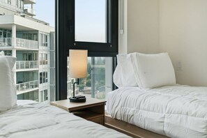 Ein Hotelzimmer mit Bett, Nachttisch, Lampe und einem Fenster mit Blick auf ein Gebäude.