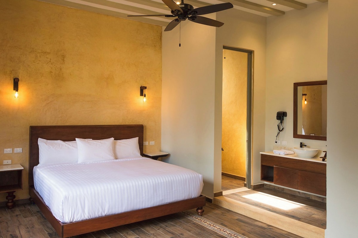 A hotel room with a bed, a ceiling fan, a bathroom with a sink and mirror, and a door leading to another room.
