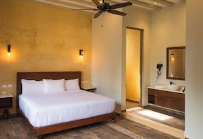 A hotel room with a bed, a ceiling fan, a bathroom with a sink and mirror, and a door leading to another room. - Hotel Casa Nuuma (Merida)