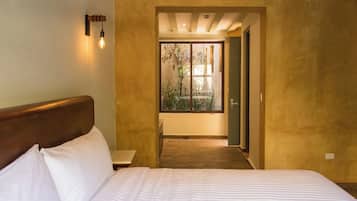 Un dormitorio con cama, mesita de noche, lámpara de pared y vistas a un área exterior con plantas a través de un vano.