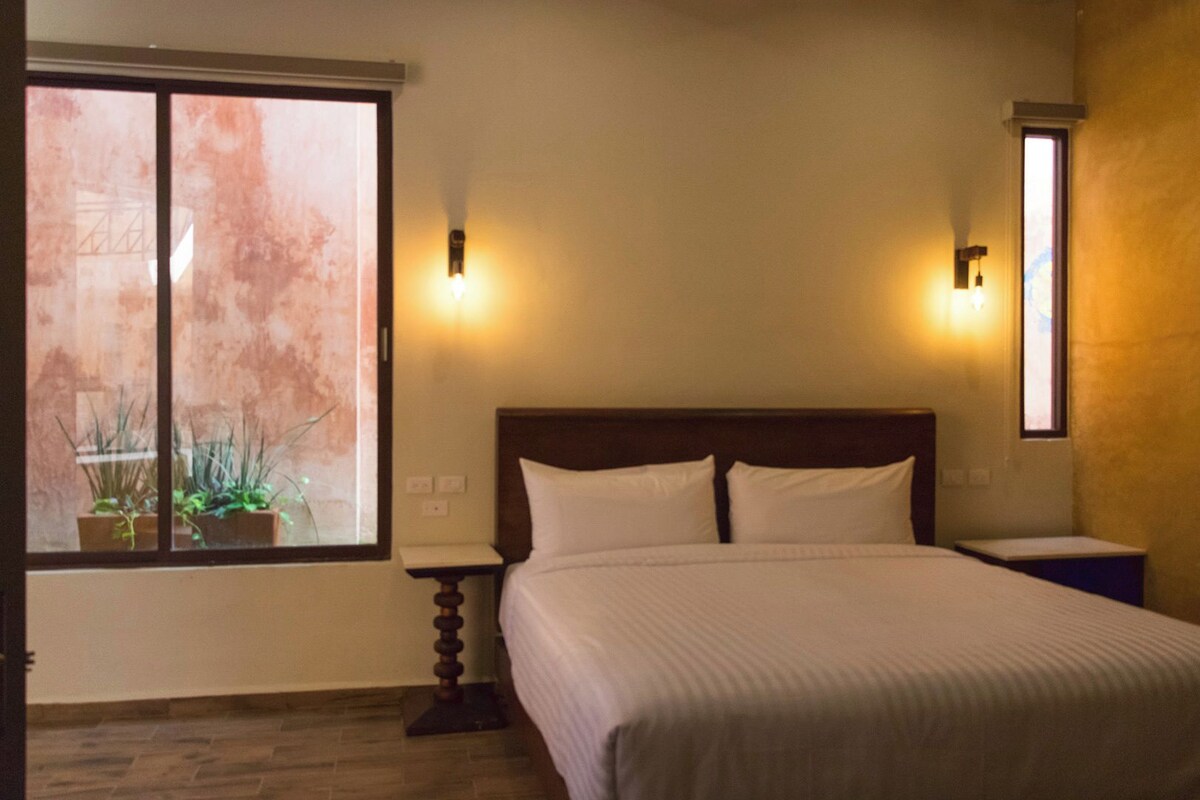 A bedroom with a large bed, two bedside lamps, a window with plants, and a view of the outside.
