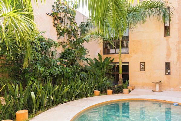 A pool area with a curved edge, surrounded by lush greenery and a building with a beige facade. - Hotel Casa Nuuma (Merida)
