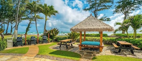 Un patio frente a la playa con un quiosco de techo de paja, mesas de picnic y estaciones de barbacoa.