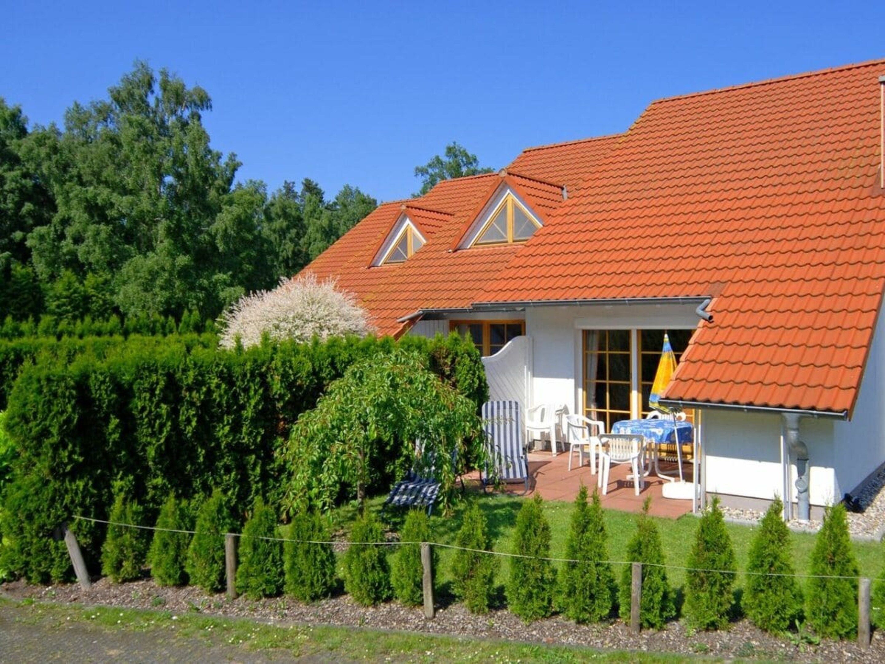 Une maison avec un toit recouvert de tuiles rouges, un petit porche avec une table et des chaises, et un jardin bien entretenu agrémenté de haies et d’arbres.