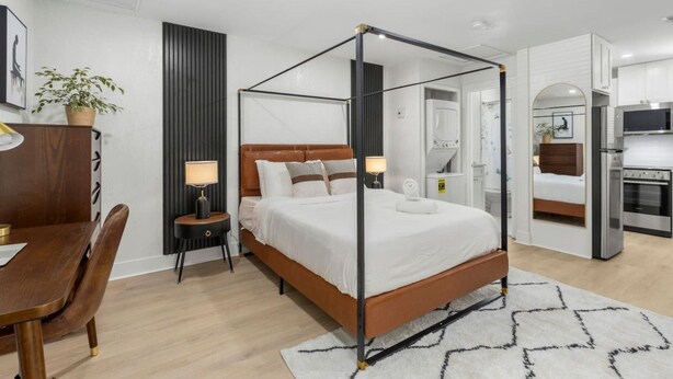 A modern bedroom with a four-poster bed, a bedside table with a lamp, a wooden desk, and a chair. - South Tampa Stay | Free Parking (Tampa)