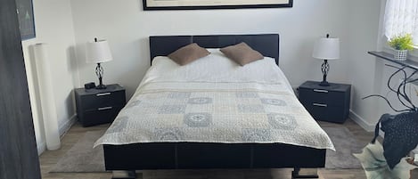 A modern bedroom with a large bed, two bedside tables, a black furry rug, a framed painting, and a window with curtains.