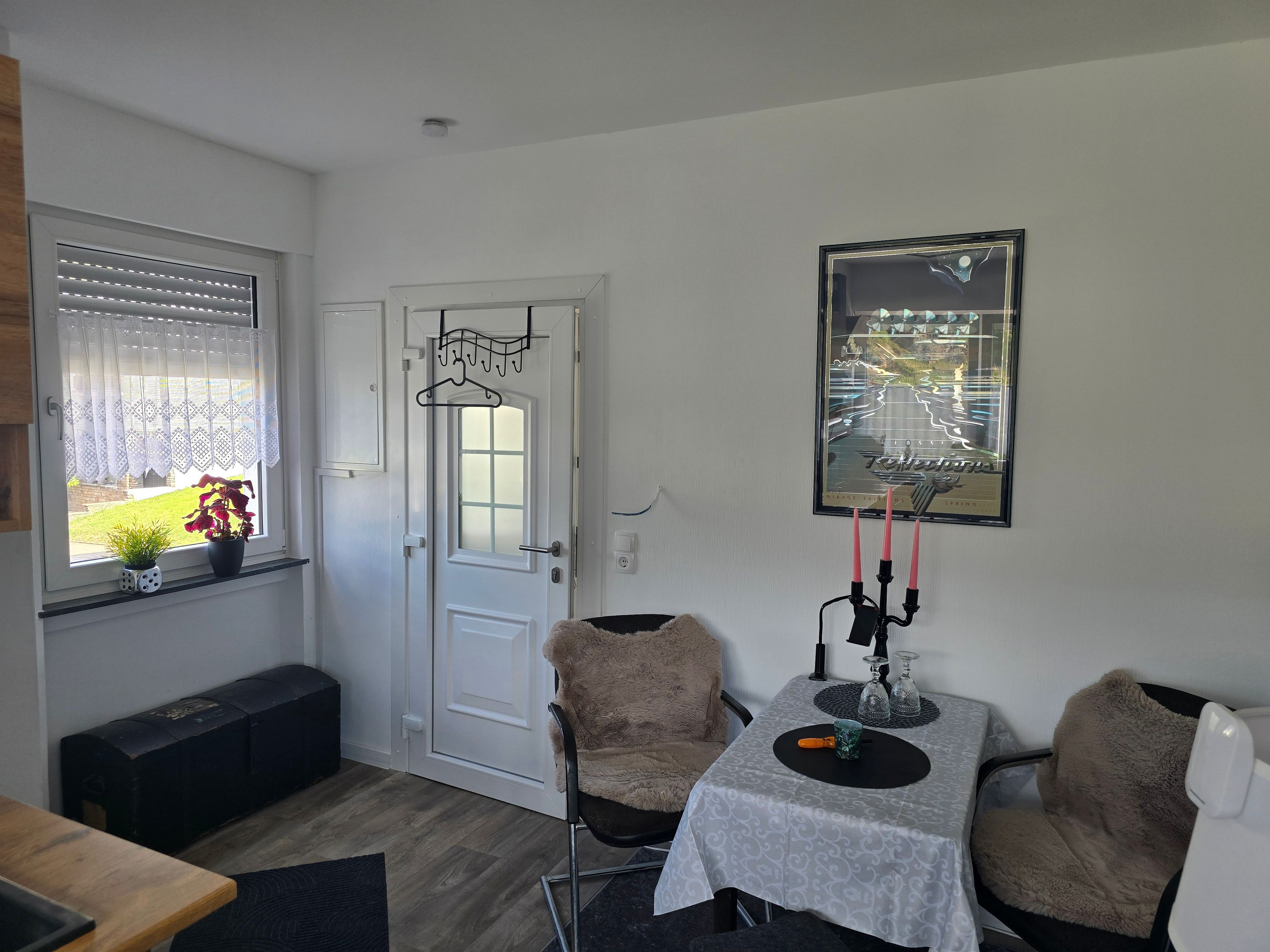 Ein Zimmer mit weißer Tür, einem Fenster mit Jalousien, einem gerahmten Poster an der Wand, einem Tisch mit Tischdecke und zwei Stühlen.