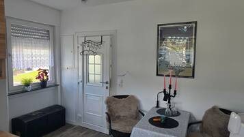 Ein Zimmer mit weißer Tür, einem Fenster mit Jalousien, einem gerahmten Poster an der Wand, einem Tisch mit Tischdecke und zwei Stühlen.
