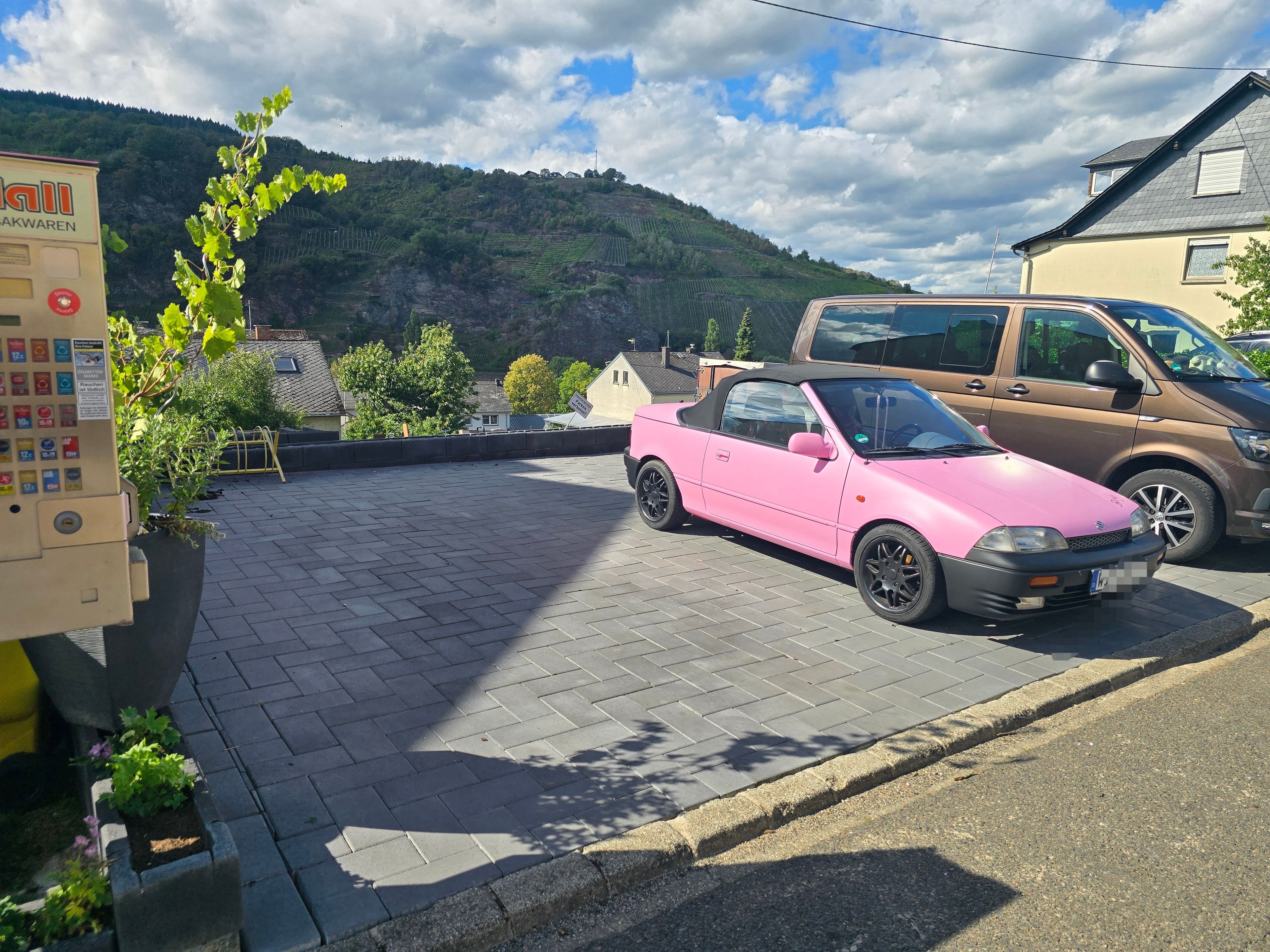 Um espaço de estacionamento com um carro conversível rosa, uma máquina de vendas automática e montanhas ao fundo.