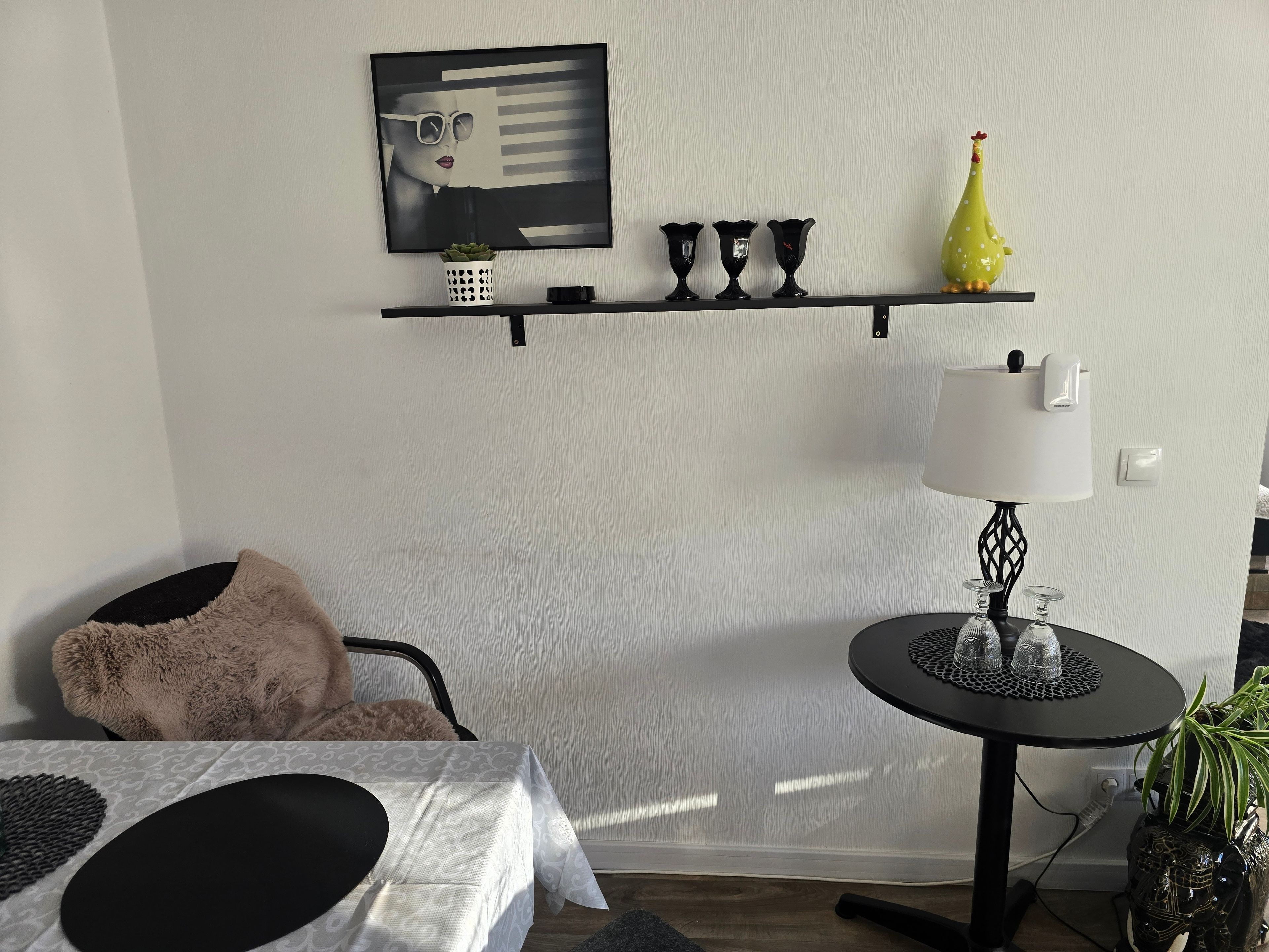 Una stanza con un letto, un tavolo con tovaglia bianca, una sedia nera, un piccolo tavolino con una lampada, una mensola a parete con oggetti decorativi e una pianta in vaso.