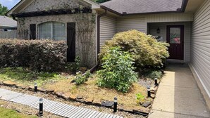 A residential house with a front porch, a garage, and a well-maintained garden with various shrubs and a wooden deck. - Cozy Cabot 3 bedroom (Cabot)