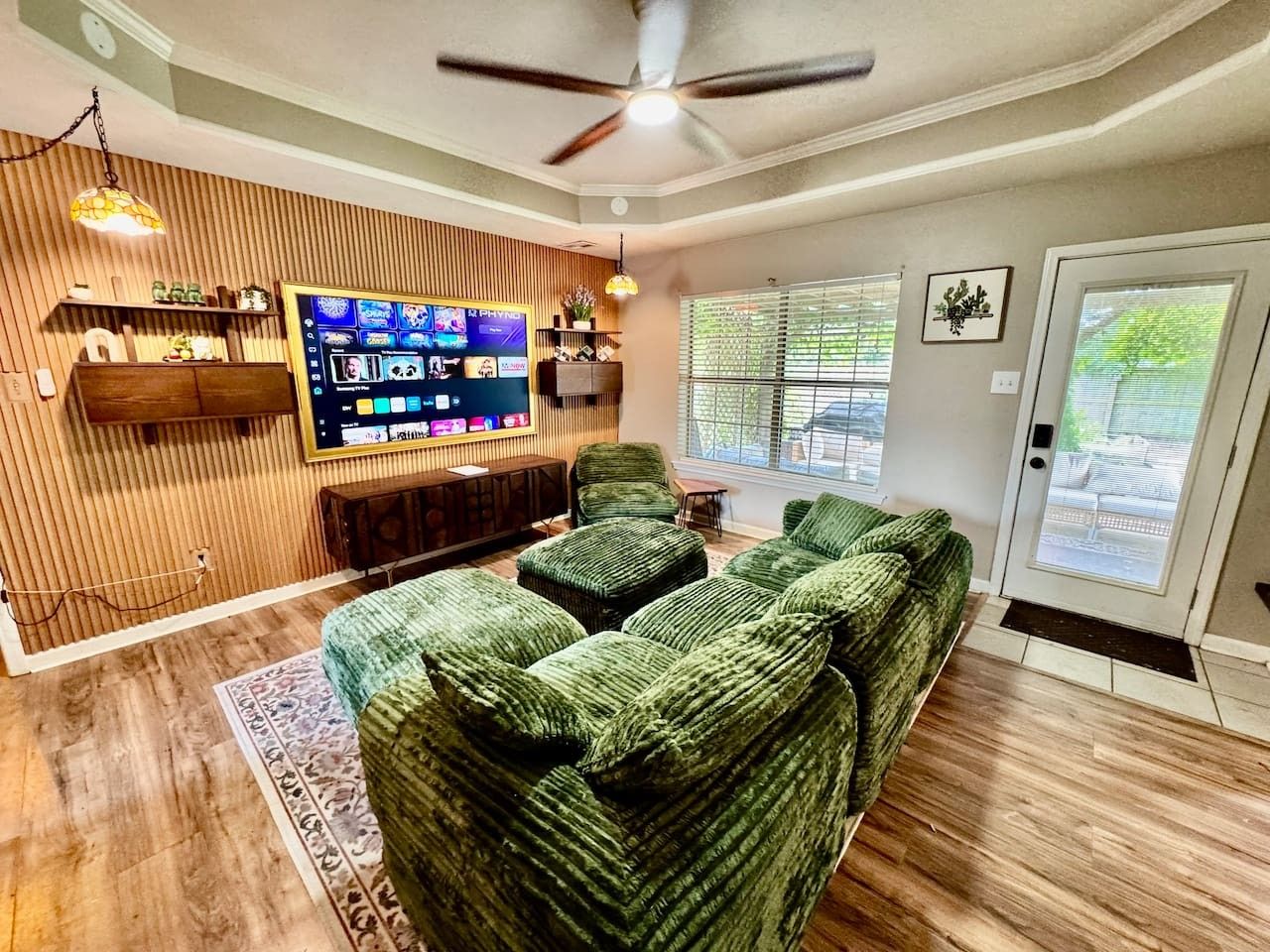 A living room with a green sofa set, a flat-screen TV mounted on a wooden wall, and a ceiling fan.