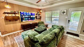 A living room with a green sofa set, a flat-screen TV mounted on a wooden wall, and a ceiling fan.