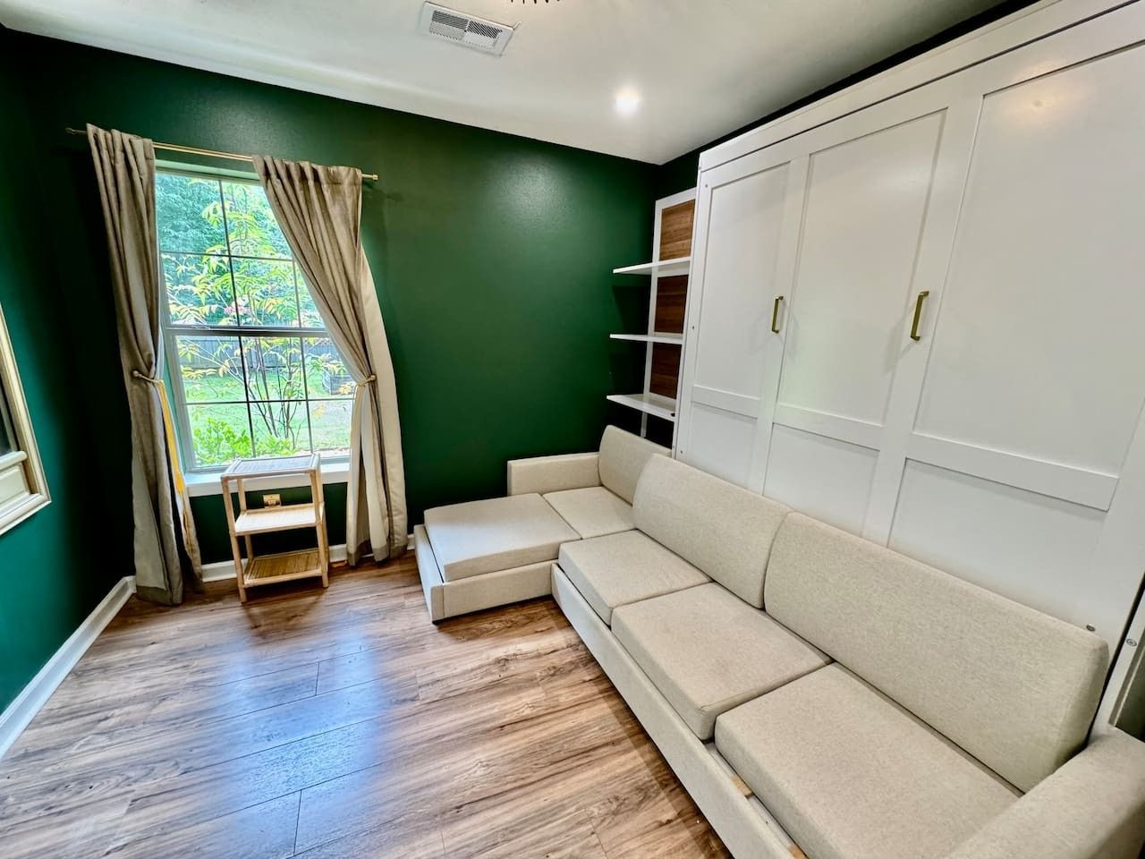 Ein Zimmer mit Ecksofa, einem kleinen Tisch, einem Kleiderschrank und einem fenster mit Vorhängen.
