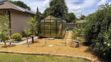 Een kas met een transparant dak, omgeven door een tuin met mulch, stapelstenen en kleine bomen.