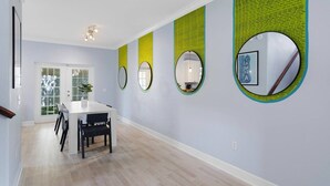Uma área de refeições com mesa e cadeiras brancas, uma porta com painéis de vidro e uma parede com três espelhos e um quadro emoldurado.
