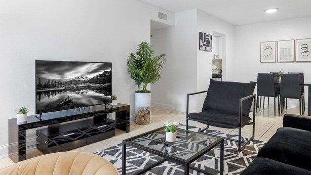 A modern living room with a flat-screen TV, a sofa, a coffee table, and a dining area with chairs. - Walk to Bayshore | Pool, Gym & Parking (Tampa)