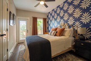 Une chambre à coucher comprenant un lit, une table de chevet, une lampe, un ventilateur de plafond et une vue sur l’extérieur par une porte.