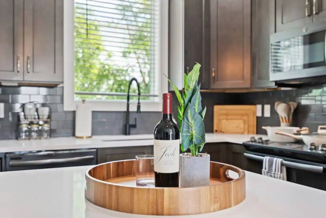Una cocina con armarios oscuros, un microondas y una botella de vino sobre una bandeja de madera.