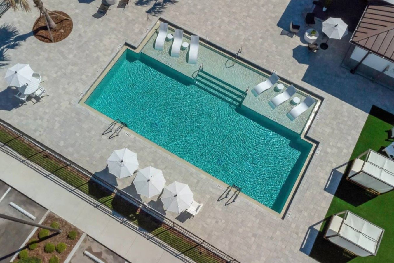 An aerial view of a swimming pool with sun loungers and umbrellas, surrounded by a paved area and greenery.