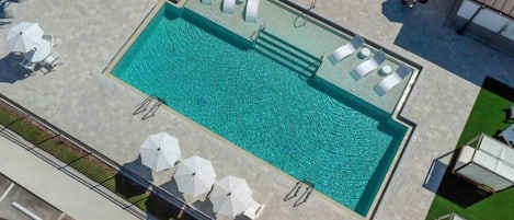 An aerial view of a swimming pool with sun loungers and umbrellas, surrounded by a paved area and greenery.