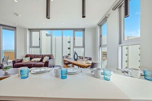 A spacious living area with a large dining table set for four, a brown leather sofa, and a view of the city through large windows. - The Reef 7C - Island Horizon (Noord)