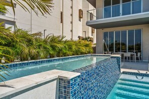 A swimming pool with blue mosaic tiles, surrounded by greenery and a building with large windows. - The Reef 7C - Island Horizon (Noord)
