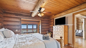 Un dormitorio tipo cabaña de madera con una cama, un aparador, un televisor y vistas a través de una ventana.